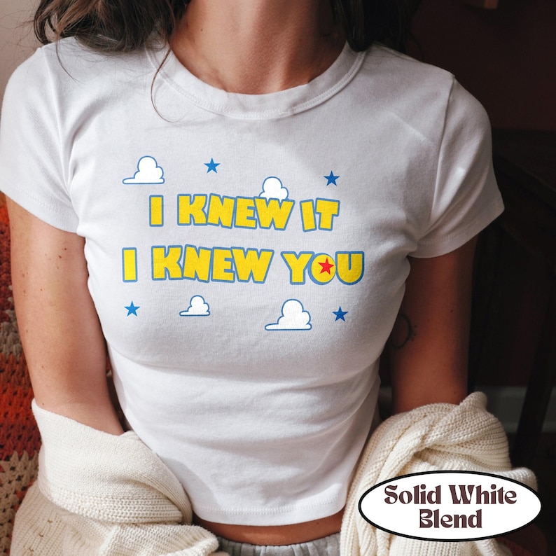 Puede incluir: Una camiseta blanca de manga corta con el texto amarillo I KNEW IT I KNEW YOU y una estrella roja dentro de la letra O. El dise&ntilde;o incluye contornos azules en el texto, peque&ntilde;as estrellas azules y nubes blancas con bordes azules al frente.