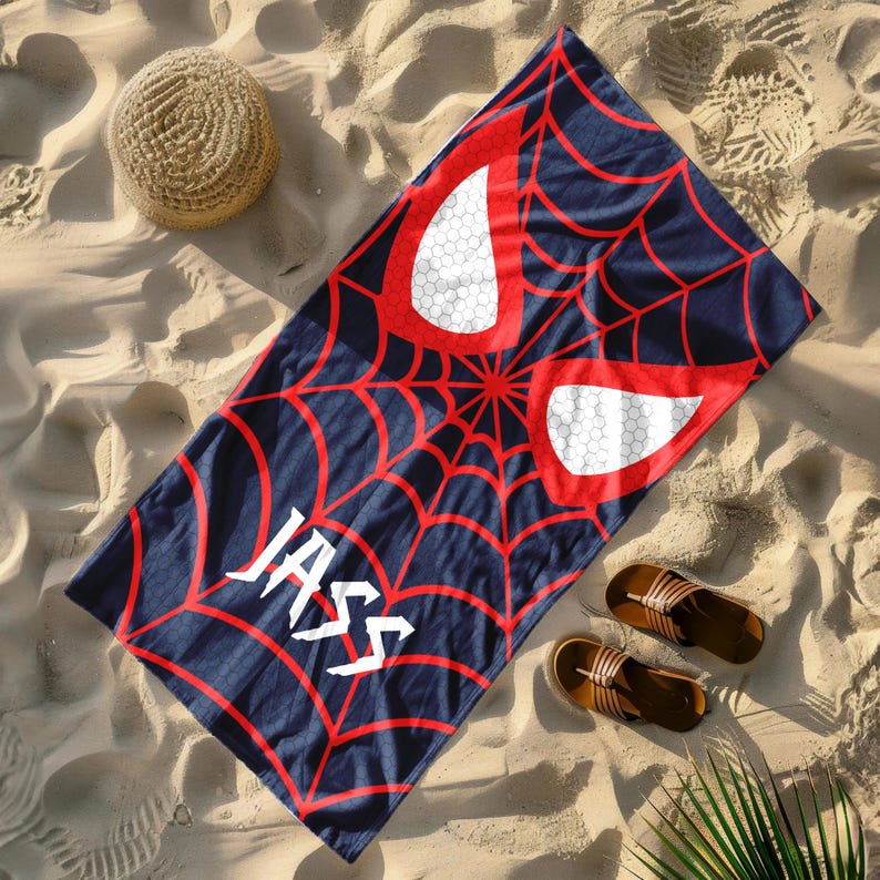 Puede incluir: Una toalla de playa azul marino con un dise&ntilde;o de telara&ntilde;a roja, formas de ojos blancos y el nombre "JASS" en blanco. Un sombrero de paja y sandalias marrones est&aacute;n en la playa arenosa.