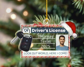 May include: A clear acrylic ornament shaped like a driver's license, featuring a photo, name, and state. The ornament includes a car key and a Santa hat. Text reads "LOOK OUT WORLD, HERE I COME!"