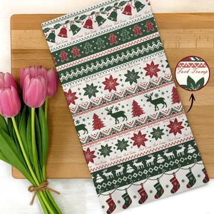 May include: A white Christmas-themed kitchen towel with a repeating pattern of red and green bells, gingerbread men, snowflakes, reindeer, and Christmas trees. The text "Fuck Trump" is visible. The towel is lying on a wooden cutting board next to a bouquet of pink tulips.