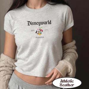 May include: Athletic heather gray t-shirt with the text "Disneyworld" in a classic font. Below the text is "EST 1971 FLORIDA" and a graphic of Disney characters. The shirt is short-sleeved.