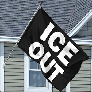May include: A black flag displaying the words "ICE OUT" in large, bold white letters. The flag is mounted on a silver pole and is displayed outside a house with grey siding and a white window.