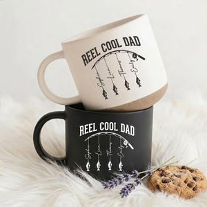 May include: Two ceramic mugs, one cream with a brown base and one matte black, both featuring the text REEL COOL DAD above a fishing rod graphic with four hanging lines, each ending in a fish icon labelled with the names Sophia, Liam, Thomas, and Emily.