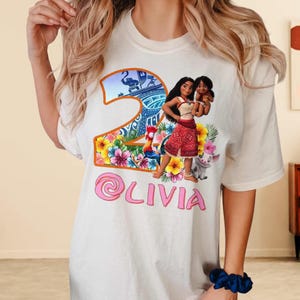 Peut inclure: T-shirt blanc avec un motif color&eacute; du chiffre 2, Moana et Pua du film Disney Moana. Le chiffre 2 est entour&eacute; de fleurs tropicales et d'un coq. Le nom Olivia est &eacute;crit en rose sous le motif.