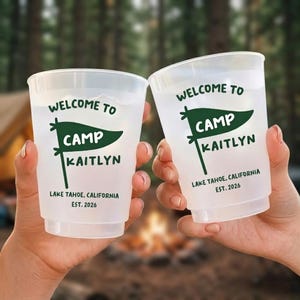 May include: Two translucent plastic cups featuring dark green text and a pennant graphic. The design reads "WELCOME TO CAMP KAITLYN" with "LAKE TAHOE, CALIFORNIA" and "EST. 2026" printed below. The cups have a simple, cylindrical shape with a smooth finish.