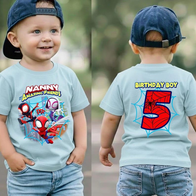 May include: A light blue t-shirt featuring a colorful graphic of Spider-Man and other characters, with the text "NANNY AMAZING FRIENDS" on the front. The back of the shirt has a large red number 5 with a spiderweb design and the words "BIRTHDAY BOY".