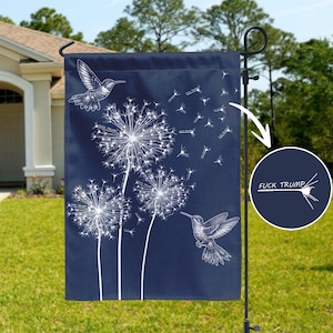 May include: A navy blue garden flag featuring white dandelion illustrations and two hummingbirds. The flag has the text "FUCK TRUMP" in a circle. The flag is hanging on a black metal pole in a yard.