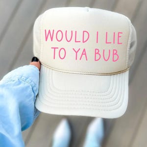 May include: A white trucker hat with a pink embroidered message that reads "Would I Lie To Ya Bub".