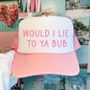 May include: A pink and white trucker hat with the phrase "WOULD I LIE TO YA BUB" in pink lettering. The hat has a pink brim and mesh backing. The hat is being held up in front of a collage of images.