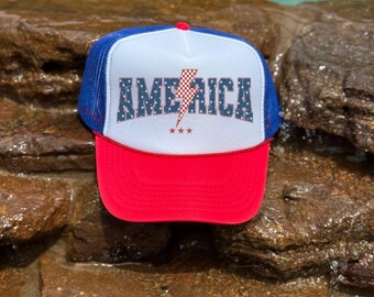 Puede incluir: Gorra de camionero roja, blanca y azul. El panel frontal muestra la palabra "AMERICA" en una fuente patriótica, un rayo y tres estrellas. La gorra tiene una visera roja, un panel frontal blanco y una parte trasera de malla azul.