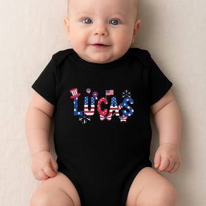 A smiling baby lies on a soft light background wearing a black personalized patriotic baby bodysuit with the custom name Lucas across the front. The name is styled in red, white, and blue with stars, stripes, fireworks, and festive patriotic accents for a bold Fourth of July look. This mockup showcases a custom baby outfit ideal for Independence Day, family holiday photos, matching sibling sets, and America’s 250th birthday celebration in 2026.