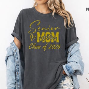 Blue jean Comfort Colors® shirt mockup with a stylish Senior Mom Class of 2026 design printed in white. The design includes elegant script lettering, large varsity MOM letters, a heart accent and personalized name placement, making it a thoughtful graduation gift for moms, mamas, bonus moms or proud senior parents supporting their 2026 graduate.