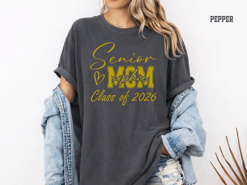 Blue jean Comfort Colors® shirt mockup with a stylish Senior Mom Class of 2026 design printed in white. The design includes elegant script lettering, large varsity MOM letters, a heart accent and personalized name placement, making it a thoughtful graduation gift for moms, mamas, bonus moms or proud senior parents supporting their 2026 graduate.