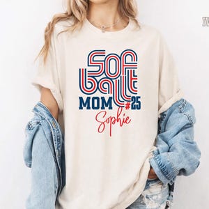 May include: Ivory-colored t-shirt with the words "SOFTBALL MOM #25 Sophie" in red and blue retro font. The shirt has a relaxed fit and is paired with a denim jacket.