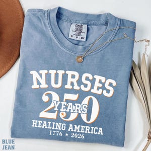 May include: A blue jean Comfort Colors t-shirt with the text "NURSES 250 YEARS HEALING AMERICA 1776 * 2026" in white and orange lettering. A gold necklace with a pendant rests on the shirt.