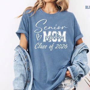 Personalized Senior Mom Comfort Colors® tee shown in blue jean with white graduation lettering for Class of 2026. This custom shirt can feature the graduate’s name across the MOM design, creating a meaningful keepsake for senior year celebrations, graduation parties, school events, senior photos, college reveal moments and proud mom memories.