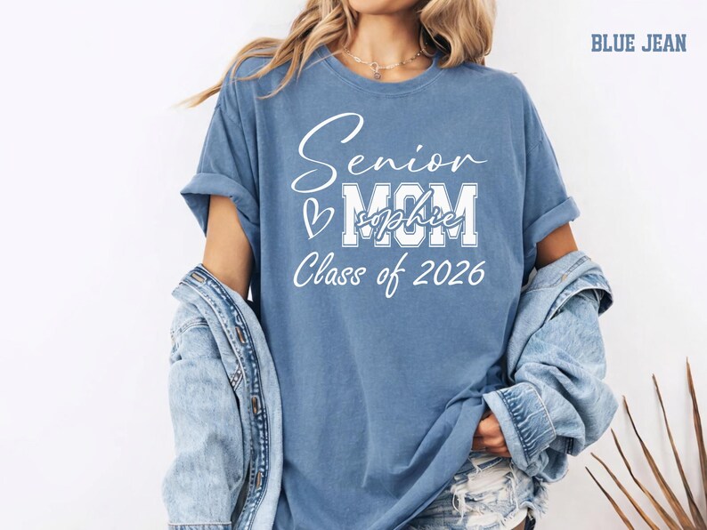 Personalized Senior Mom Comfort Colors® tee shown in blue jean with white graduation lettering for Class of 2026. This custom shirt can feature the graduate’s name across the MOM design, creating a meaningful keepsake for senior year celebrations, graduation parties, school events, senior photos, college reveal moments and proud mom memories.