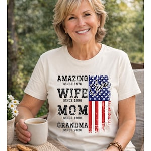 Smiling mature woman wearing a natural color t-shirt with a personalized family timeline design. The shirt features bold distressed text reading Amazing Since, Wife, Mom, Grandma with custom years, alongside a vertical American flag graphic. Perfect for patriotic celebrations like Fourth of July or meaningful gifts for Mother’s Day. The relaxed outdoor setting and warm tones highlight the comfort and emotional value of this custom grandma shirt.