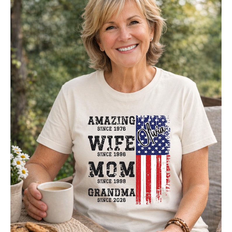 Smiling mature woman wearing a natural color t-shirt with a personalized family timeline design. The shirt features bold distressed text reading Amazing Since, Wife, Mom, Grandma with custom years, alongside a vertical American flag graphic. Perfect for patriotic celebrations like Fourth of July or meaningful gifts for Mother’s Day. The relaxed outdoor setting and warm tones highlight the comfort and emotional value of this custom grandma shirt.