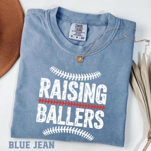 This Raising Ballers Comfort Colors® shirt is perfect for proud baseball and softball moms who spend their days cheering at the ballpark. Featuring a vintage style baseball graphic, this soft garment dyed tee is great for game days, tournaments, and everyday casual wear. A must have for sports moms who proudly support their young athletes and love the ballpark lifestyle.
