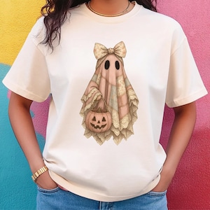 May include: Cream-colored t-shirt featuring a whimsical ghost design. The ghost is draped in a patchwork quilt with a bow and holds a jack-o'-lantern. The design incorporates soft pinks, creams, and beige tones, perfect for Halloween.
