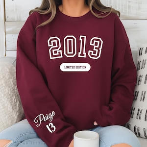 A person wearing a burgundy sweatshirt with the year "2013" printed on it, along with the text "LIMITED EDITION" and the name " Paige 13".