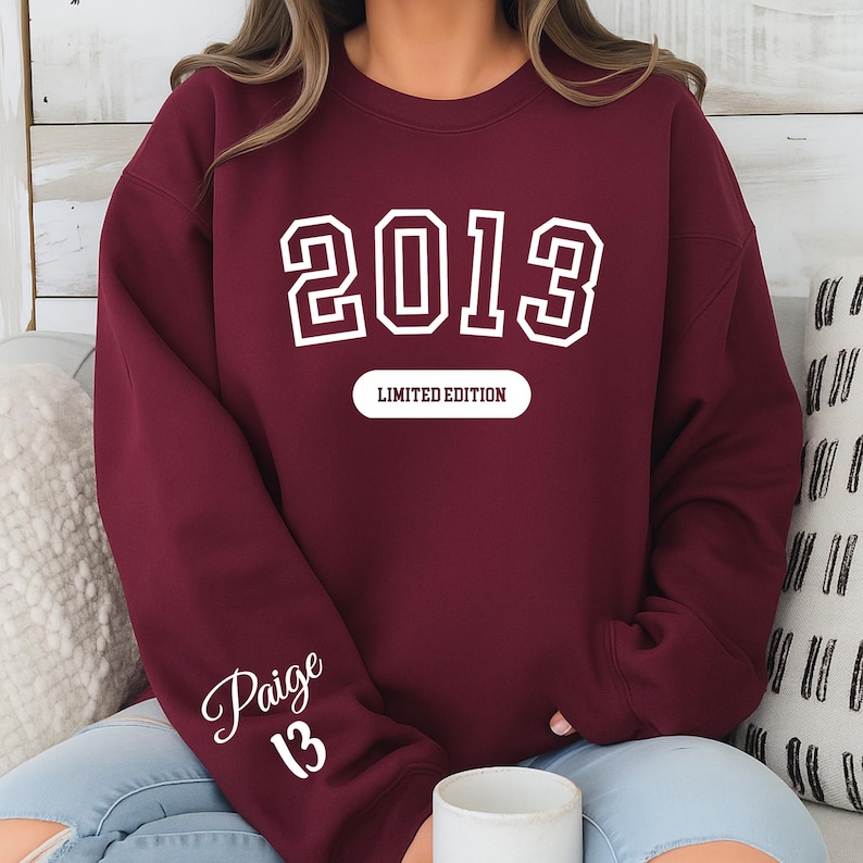A person wearing a burgundy sweatshirt with the year "2013" printed on it, along with the text "LIMITED EDITION" and the name " Paige 13".