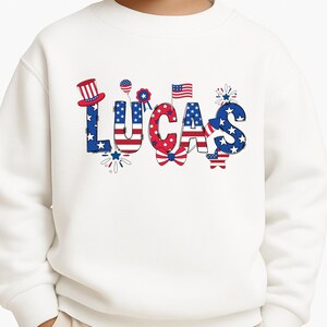 A close-up mockup shows the front of a white kids sweatshirt with the personalized name Lucas printed in large patriotic lettering. The custom design combines red, white, and blue colors, stars, stripes, fireworks, and festive Americana details that make it ideal for Independence Day and the 250th anniversary of the United States. This image clearly highlights the print placement, design scale, and personalized name option for boys or girls.
