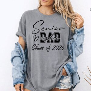 Blue jean Comfort Colors® graduation mom shirt with white printed lettering for Class of 2026. The design combines script Senior text, varsity MOM letters, heart accent and custom name placement for a trendy personalized look. A great gift for moms celebrating their child’s senior year, graduation party, senior photos and school milestones.