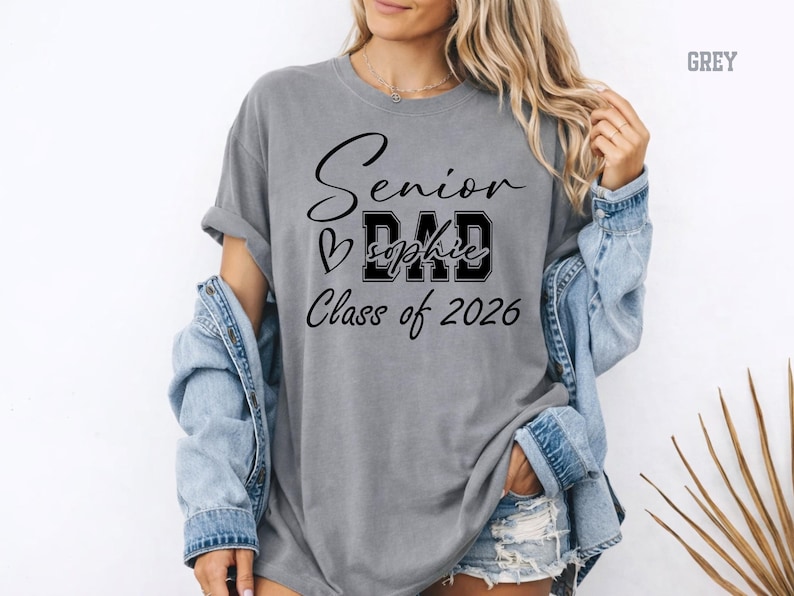 Blue jean Comfort Colors® graduation mom shirt with white printed lettering for Class of 2026. The design combines script Senior text, varsity MOM letters, heart accent and custom name placement for a trendy personalized look. A great gift for moms celebrating their child’s senior year, graduation party, senior photos and school milestones.