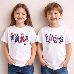 Two smiling young children stand side by side wearing matching personalized patriotic shirts in a soft natural cream color. One shirt features the custom name Lucas and the other shows the custom name Emma in bold red, white, and blue lettering with stars, stripes, fireworks, a patriotic bow, and festive USA-inspired details. This personalized kids Fourth of July shirt mockup highlights a custom name design created for Independence Day and America’s 250th anniversary celebration.