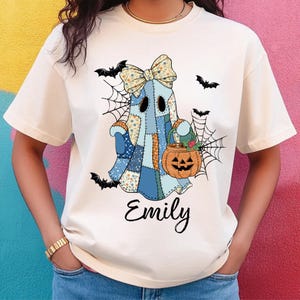 May include: Cream-colored t-shirt featuring a patchwork ghost design with a bow, holding a pumpkin basket. The ghost is surrounded by bats and spiderwebs. The name "Emily" is written in cursive below the ghost.