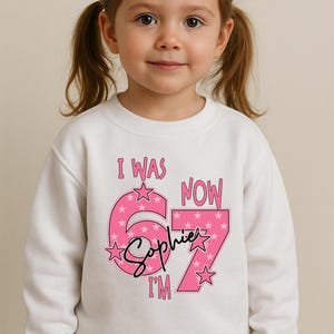 A young girl wearing a white sweatshirt with a pink and purple design and text on it.