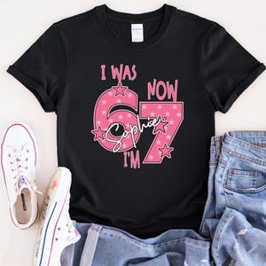 A black t-shirt with a pink and white design that says "I WAS NOW 67" and "Sophie I'M" printed on it. The t-shirt is placed on top of a pair of jeans and white sneakers.