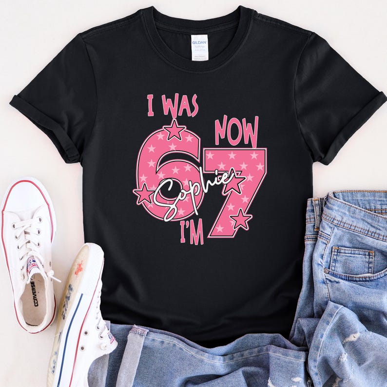 A black t-shirt with a pink and white design that says "I WAS NOW 67" and "Sophie I'M" printed on it. The t-shirt is placed on top of a pair of jeans and white sneakers.