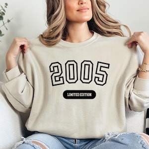 A woman wearing a beige sweater with the number "2005" printed on it, posing with her hands on her shoulders.