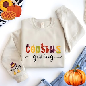 May include: Cream-colored sweatshirt with the words "Cousins giving" in colorful letters. The word "Cousins" has Thanksgiving-themed icons in place of the letter "O" and "S". The left sleeve has a pilgrim hat and the name "Emily".