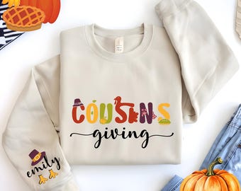 May include: Cream-colored sweatshirt with the words "Cousins giving" in colorful letters. The word "Cousins" has Thanksgiving-themed icons in place of the letter "O" and "S". The left sleeve has a pilgrim hat and the name "Emily".