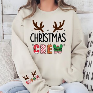 cute christmas crew sweatshirt with reindeer antlers and personalized sleeve name option, matching holiday family sweater for kids and adults, custom christmas shirt for winter celebrations, cozy xmas crew top with festive lettering, personalized holiday gift sweatshirt, christmas pajamas top for family photos, fun reindeer themed christmas sweater, holiday party outfit idea, custom name christmas crew shirt, adorable winter sweatshirt for holiday season
