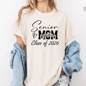 Personalized Comfort Colors® senior mom shirt featuring Class of 2026 text, large MOM lettering, handwritten style Senior script and a custom graduate name across the center. Shown on a blue jean tee with white print, this graduation mom shirt is perfect for proud senior moms, school events, graduation gifts and Class of 2026 memories.
