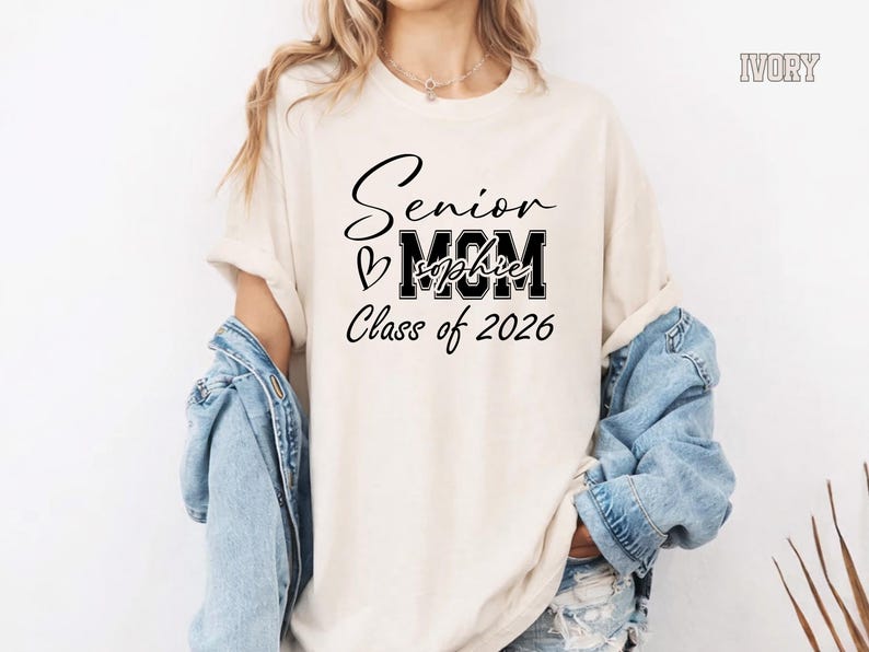 Personalized Comfort Colors® senior mom shirt featuring Class of 2026 text, large MOM lettering, handwritten style Senior script and a custom graduate name across the center. Shown on a blue jean tee with white print, this graduation mom shirt is perfect for proud senior moms, school events, graduation gifts and Class of 2026 memories.