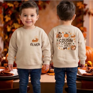 May include: A beige crewneck sweatshirt featuring a graphic of a slice of pumpkin pie and the name Oliver on the front left chest. The back displays a turkey, pumpkin, pie, mug, and basket with the text Cousin Crew 2026 in brown lettering.