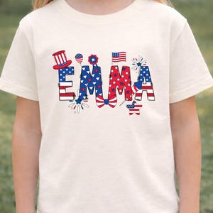 A front-facing outdoor mockup shows a child wearing a natural cream personalized patriotic T-shirt with the custom name Emma printed across the chest. The design features red, white, and blue colors, stars, stripes, fireworks, and festive Americana elements for a playful holiday look. This custom kids shirt is designed for Fourth of July celebrations, matching family outfits, summer events, and the 250th anniversary of America in 2026.