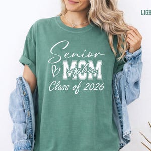 Proud senior mom Comfort Colors® shirt featuring a white varsity and script design on a blue jean tee. The personalized graduate name across the MOM lettering adds a special custom touch for Class of 2026 celebrations. Ideal for graduation gifts, senior parent outfits, mom photo shoots, school events and everyday senior year pride.