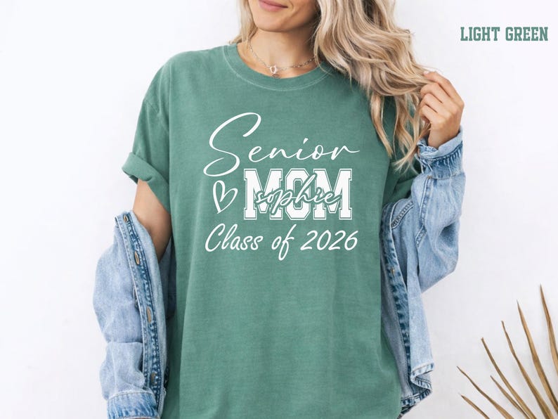 Proud senior mom Comfort Colors® shirt featuring a white varsity and script design on a blue jean tee. The personalized graduate name across the MOM lettering adds a special custom touch for Class of 2026 celebrations. Ideal for graduation gifts, senior parent outfits, mom photo shoots, school events and everyday senior year pride.