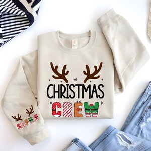 cute christmas crew sweatshirt with reindeer antlers and personalized sleeve name option, matching holiday family sweater for kids and adults, custom christmas shirt for winter celebrations, cozy xmas crew top with festive lettering, personalized holiday gift sweatshirt, christmas pajamas top for family photos, fun reindeer themed christmas sweater, holiday party outfit idea, custom name christmas crew shirt, adorable winter sweatshirt for holiday season