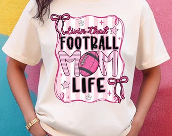 May include: A cream-colored t-shirt with a pink and white graphic that reads "Livin that FOOTBALL MOM LIFE." The graphic includes a football, stars, and bows. The shirt has short sleeves and a crew neck.
