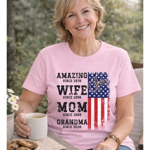 Outdoor lifestyle mockup of a happy older woman holding a coffee cup while wearing a custom grandma t-shirt. The design includes personalized years for wife, mom, and grandma roles, paired with an American flag graphic. This shirt is perfect for proud grandmothers who want to showcase their family journey with style. Suitable for casual wear, holidays, and patriotic events, combining comfort with meaningful personalization.