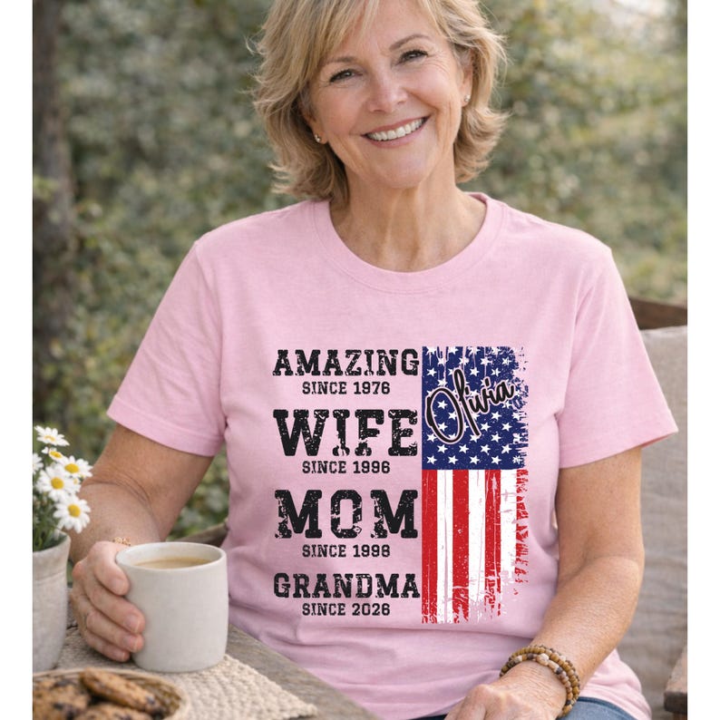 Outdoor lifestyle mockup of a happy older woman holding a coffee cup while wearing a custom grandma t-shirt. The design includes personalized years for wife, mom, and grandma roles, paired with an American flag graphic. This shirt is perfect for proud grandmothers who want to showcase their family journey with style. Suitable for casual wear, holidays, and patriotic events, combining comfort with meaningful personalization.