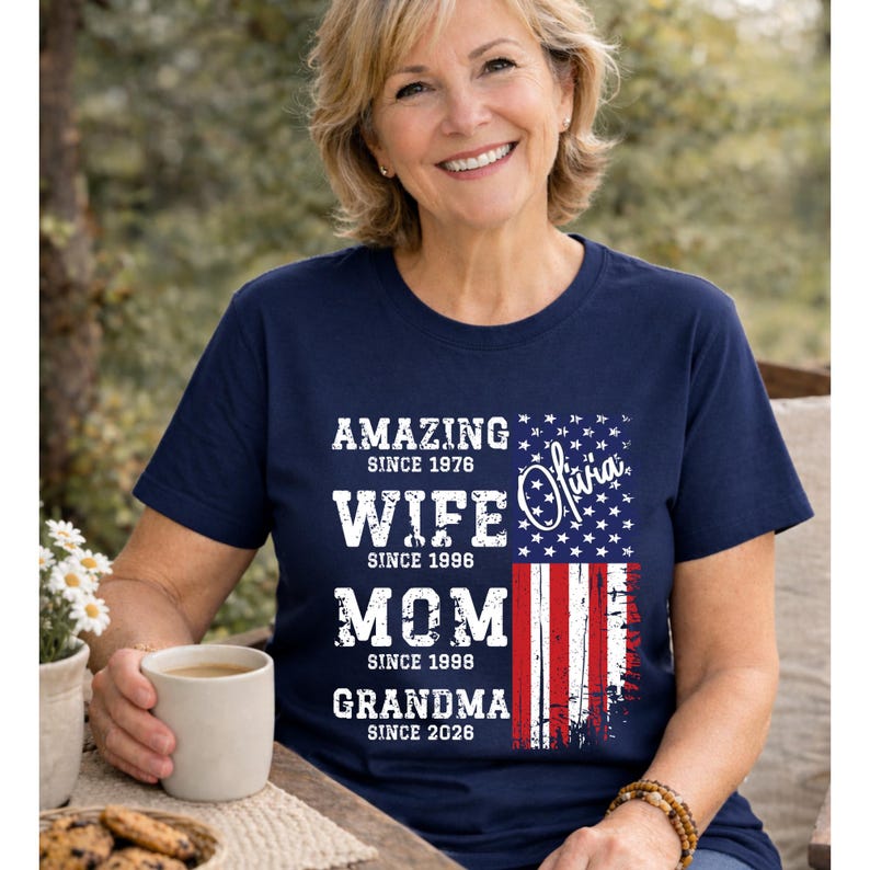 Natural color t-shirt featuring a personalized family role timeline design with bold black typography and a distressed American flag print. The text highlights milestones like wife, mom, and grandma with custom dates, making it a unique and sentimental gift. Styled on a smiling woman in a cozy outdoor setting, this shirt is ideal for Mother’s Day, grandma gifts, or celebrating family pride in a stylish and patriotic way.